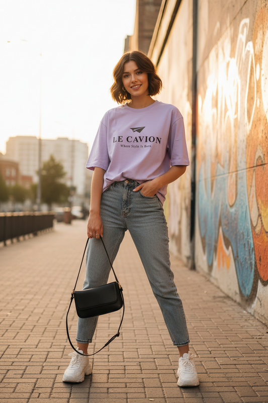 Woman wearing a light purple t-shirt with text, blue jeans, and white sneakers, standing on a sidewalk with graffiti in the background.