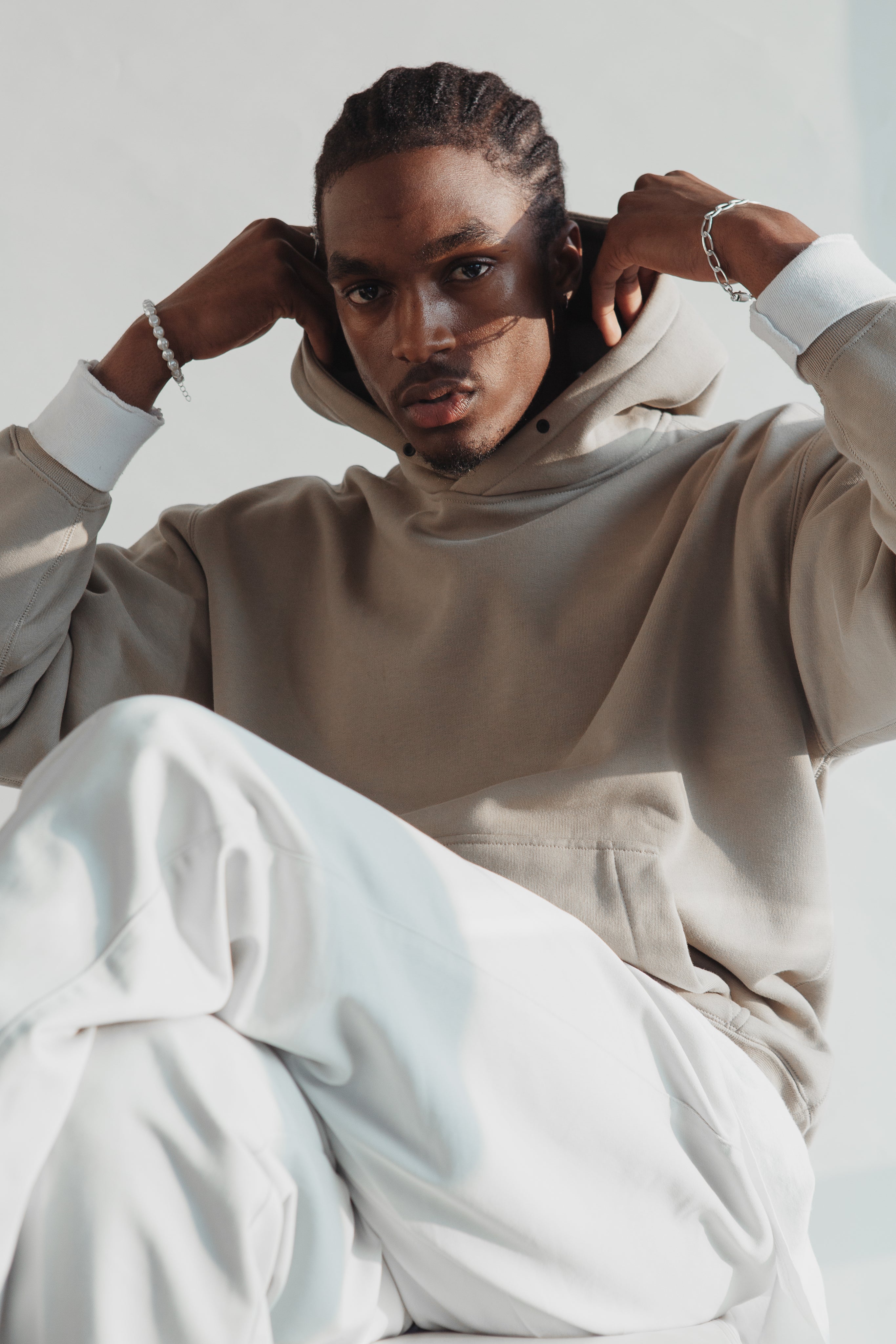 A person with braided hair sits casually while adjusting the hood of a beige hoodie. They are wearing loose white pants and simple jewelry, including bracelets. Soft natural light casts gentle shadows across their face against a clean, minimal background.